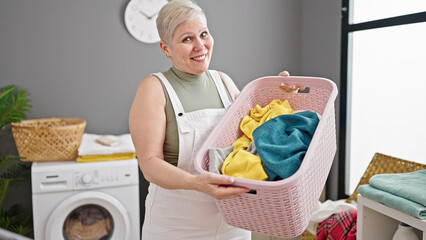 Wall Mural - Middle age grey-haired woman smiling confident holding basket with clothes at laundry room