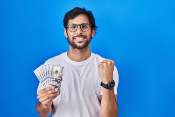 Wall Mural - Handsome latin man holding dollars banknotes screaming proud, celebrating victory and success very excited with raised arms