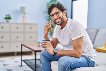 Wall Mural - Young hispanic man drinking coffee and talking on the smartphone at home