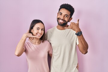 Poster - Young hispanic couple together over pink background smiling doing phone gesture with hand and fingers like talking on the telephone. communicating concepts.