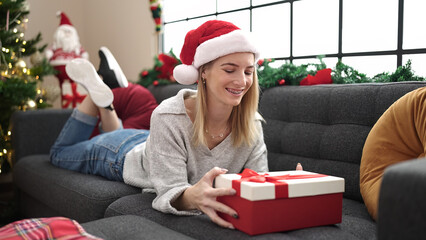 Poster - Young blonde woman unpacking gift lying on the sofa by christmas tree at home