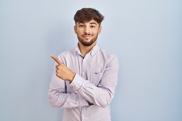 Wall Mural - Arab man with beard standing over blue background pointing with hand finger to the side showing advertisement, serious and calm face