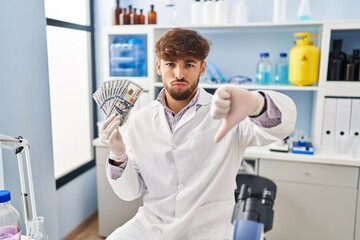 Canvas Print - Arab man with beard working at scientist laboratory holding money with angry face, negative sign showing dislike with thumbs down, rejection concept