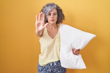 Middle age woman with grey hair wearing pijama hugging pillow doing stop sing with palm of the hand. warning expression with negative and serious gesture on the face.