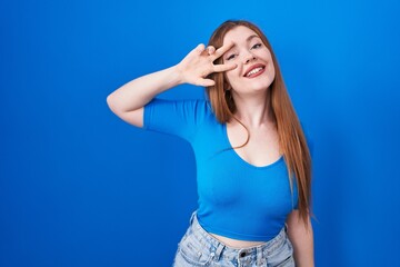 Sticker - Redhead woman standing over blue background doing peace symbol with fingers over face, smiling cheerful showing victory