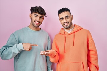 Canvas Print - Young hispanic gay couple standing over pink background amazed and smiling to the camera while presenting with hand and pointing with finger.