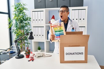 Canvas Print - Hispanic young doctor man donating toys at the clinic angry and mad screaming frustrated and furious, shouting with anger. rage and aggressive concept.