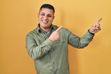 Canvas Print - Hispanic young man standing over yellow background smiling and looking at the camera pointing with two hands and fingers to the side.