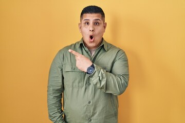 Canvas Print - Hispanic young man standing over yellow background surprised pointing with finger to the side, open mouth amazed expression.