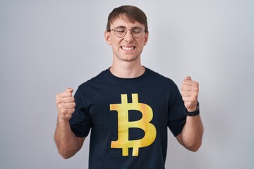 Canvas Print - Caucasian blond man wearing bitcoin t shirt excited for success with arms raised and eyes closed celebrating victory smiling. winner concept.