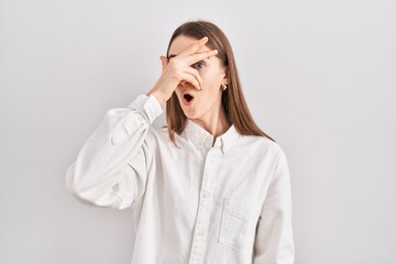 Sticker - Young caucasian woman standing over isolated background peeking in shock covering face and eyes with hand, looking through fingers with embarrassed expression.