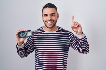 Wall Mural - Young hispanic man wearing pyjama holding alarm clock surprised with an idea or question pointing finger with happy face, number one