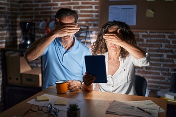 Sticker - Middle age hispanic couple using touchpad sitting on the table at night covering eyes with hand, looking serious and sad. sightless, hiding and rejection concept