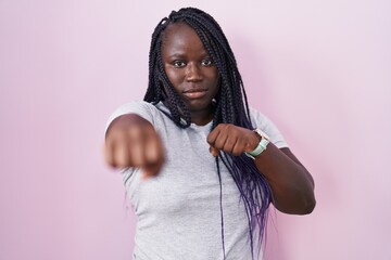 Sticker - Young african woman standing over pink background punching fist to fight, aggressive and angry attack, threat and violence