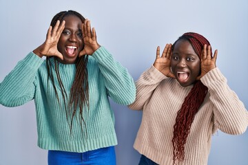 Canvas Print - Two african woman standing over blue background smiling cheerful playing peek a boo with hands showing face. surprised and exited