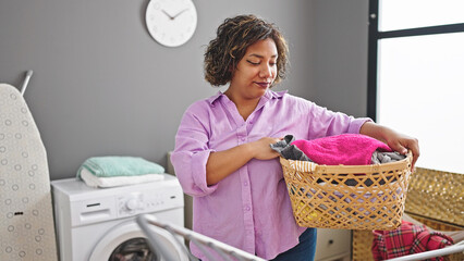 Wall Mural - Young beautiful latin woman holding basket with clothes with serious face at laundry room