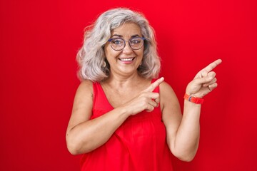 Poster - Middle age woman with grey hair standing over red background smiling and looking at the camera pointing with two hands and fingers to the side.