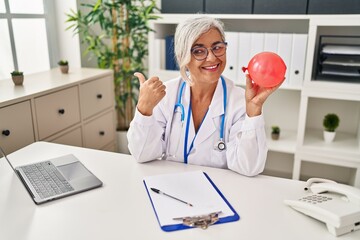 Poster - Middle age woman with grey hair wearing doctor uniform holding balloon pointing thumb up to the side smiling happy with open mouth