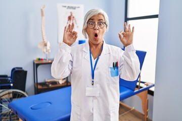 Poster - Middle age woman with grey hair working at pain recovery clinic looking surprised and shocked doing ok approval symbol with fingers. crazy expression