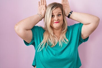 Poster - Caucasian plus size woman standing over pink background doing bunny ears gesture with hands palms looking cynical and skeptical. easter rabbit concept.