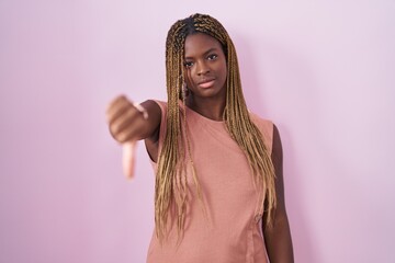 Wall Mural - African american woman with braided hair standing over pink background looking unhappy and angry showing rejection and negative with thumbs down gesture. bad expression.