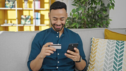 Wall Mural - Young hispanic man doing online shopping sitting on sofa at home