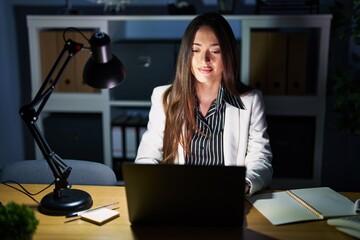 Poster - Young brunette woman working at the office at night with laptop relaxed with serious expression on face. simple and natural looking at the camera.