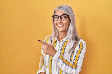 Wall Mural - Middle age woman with grey hair standing over yellow background wearing glasses cheerful with a smile of face pointing with hand and finger up to the side with happy and natural expression on face