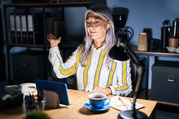 Poster - Middle age woman with grey hair working at the office at night smiling cheerful presenting and pointing with palm of hand looking at the camera.