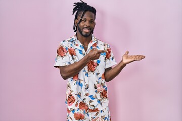 Sticker - African man with dreadlocks wearing summer shirt over pink background amazed and smiling to the camera while presenting with hand and pointing with finger.