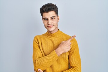 Wall Mural - Young hispanic man standing over blue background pointing with hand finger to the side showing advertisement, serious and calm face