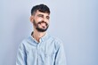 © Krakenimages.com - Young hispanic man with beard standing over blue background looking away to side with smile on face, natural expression. laughing confident.