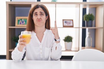 Wall Mural - Brunette woman drinking glass of orange juice pointing up looking sad and upset, indicating direction with fingers, unhappy and depressed.