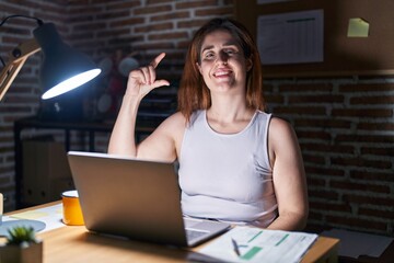 Sticker - Brunette woman working at the office at night smiling and confident gesturing with hand doing small size sign with fingers looking and the camera. measure concept.