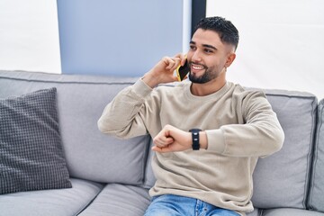 Wall Mural - Young arab man talking on the smartphone looking watch at home