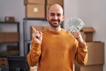 Canvas Print - Young bald man with beard working at small business ecommerce holding money smiling happy pointing with hand and finger to the side