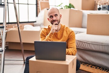 Young bald man with beard moving to a new house using laptop serious face thinking about question with hand on chin, thoughtful about confusing idea