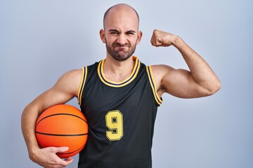 Poster - Young bald man with beard wearing basketball uniform holding ball strong person showing arm muscle, confident and proud of power