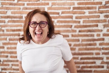 Sticker - Senior woman with glasses standing over bricks wall angry and mad screaming frustrated and furious, shouting with anger. rage and aggressive concept.