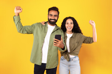 Wall Mural - Arab couple celebrating holding smarphone and shaking fists, yellow background