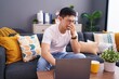 Young asian man using laptop at home sitting on the sofa smelling something stinky and disgusting, intolerable smell, holding breath with fingers on nose. bad smell