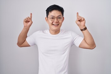 Sticker - Young asian man standing over white background smiling amazed and surprised and pointing up with fingers and raised arms.