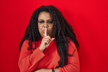 Poster - Plus size hispanic woman standing over red background asking to be quiet with finger on lips. silence and secret concept.