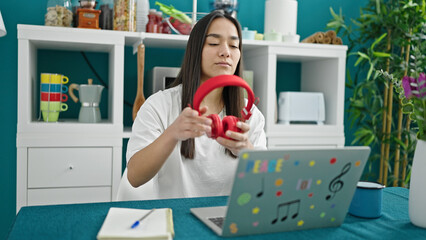 Poster - Young beautiful hispanic woman using laptop and headphones sitting on table at dinning room