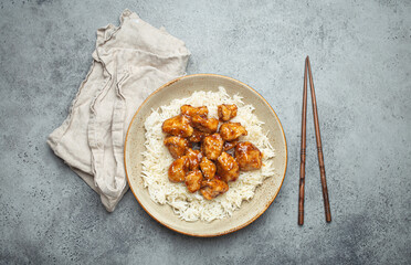 Poster - Chinese sweet and sour sticky chicken with sesame seeds and rice on ceramic plate with chopsticks top view on gray rustic stone background, traditional dish of China