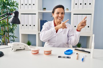 Sticker - Hispanic mature dentist woman working at the clinic smiling and looking at the camera pointing with two hands and fingers to the side.