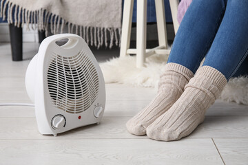 Wall Mural - Woman in warm socks near electric fan heater at home, closeup