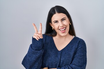 Poster - Young brunette woman standing over isolated background smiling with happy face winking at the camera doing victory sign with fingers. number two.