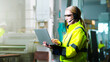 © ihorvsn - Caucasian female young engineer in headset and yellow uniform standing in factory, talking with video and working at laptop. Engineering concept. Indoor. Woman videochatting on computer at plant.