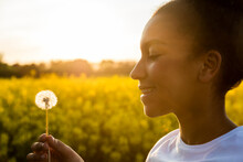 Outdoor Portrait Of Beautiful Happy Mixed Race African American Girl Teenager Female Young Woman Smiling In Golden Sunset Light Blowing Dandelion Clock Flower